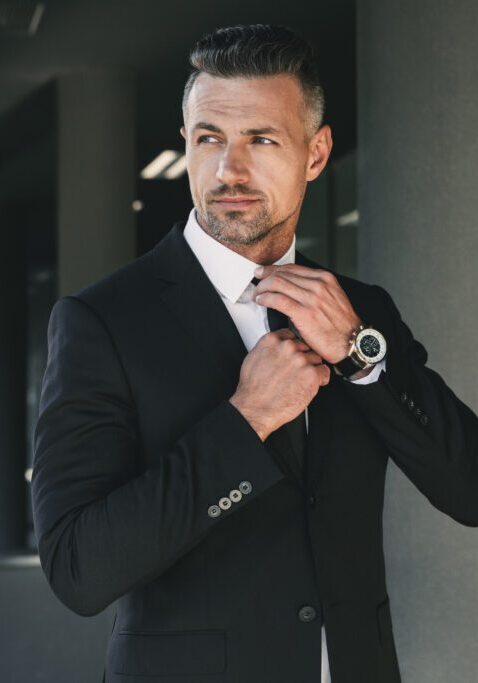 Portrait of a charming young businessman dressed in suit standing outside a glass building and touching his tie